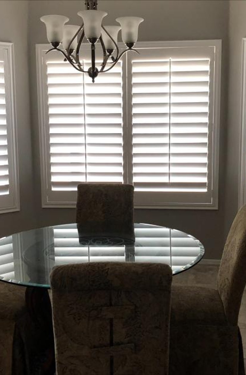 white plantation shutters across a bay window with a round glass dining table